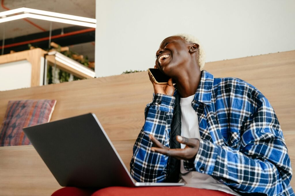 Cheerful woman in checkered shirt using smartphone and laptop indoors, embodying freelance lifestyle.